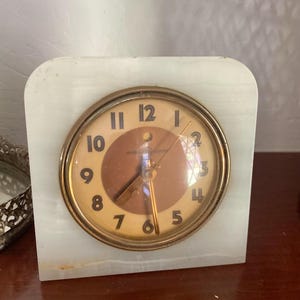 May include: A vintage white marble clock with a brass face and black numbers. The clock face has a gold-colored hour and minute hand and a red second hand. The clock face reads "General Electric".