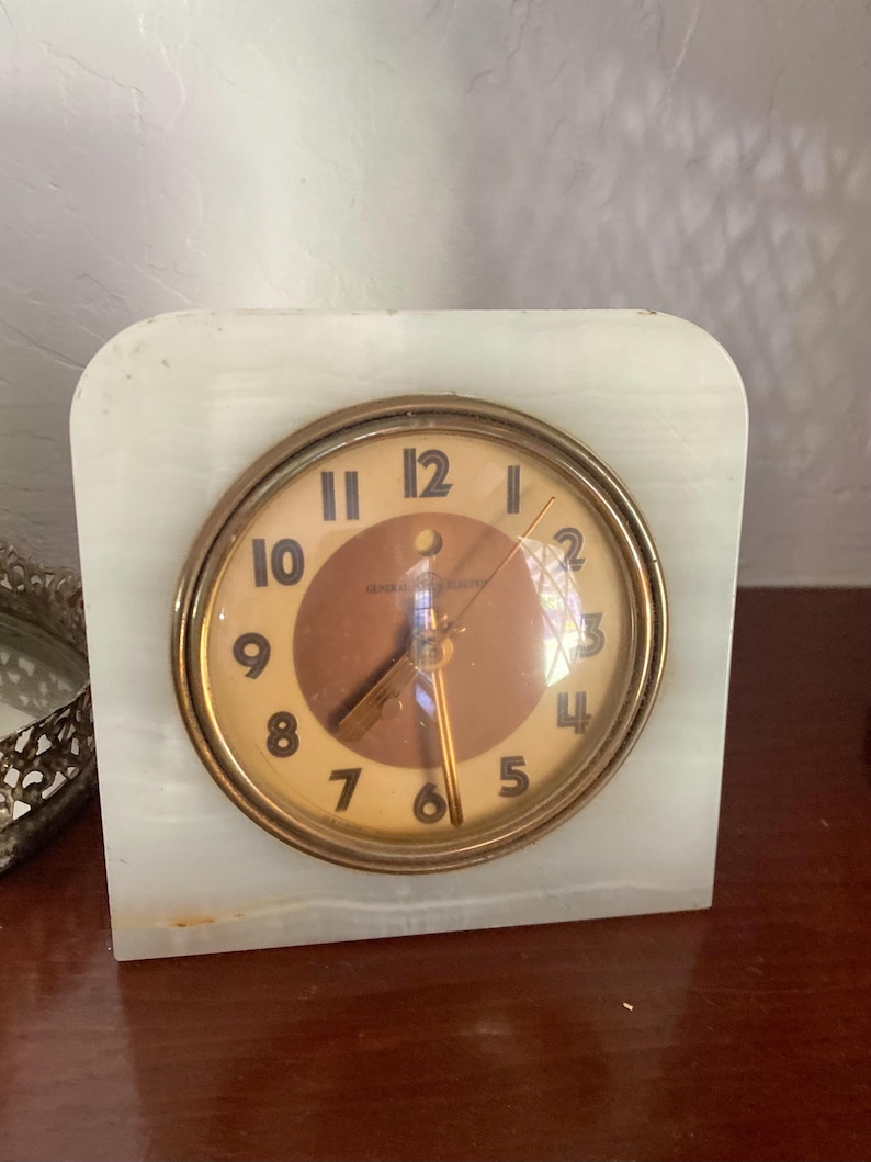 May include: A vintage white marble clock with a brass face and black numbers. The clock face has a gold-colored hour and minute hand and a red second hand. The clock face reads "General Electric".