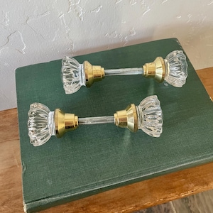May include: Two door knobs featuring clear, fluted glass handles with gold-toned metal bases and a central threaded metal spindle connecting the two ends. The design includes a faceted glass surface and a polished metallic finish on the hardware.