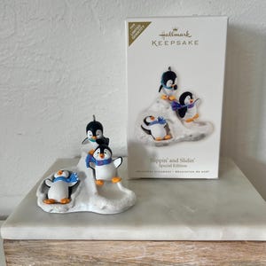 Peut inclure: Ornement de Noël Hallmark avec trois pingouins sur une colline enneigée. Les pingouins noirs, blancs et oranges portent des écharpes bleues et violettes. La boîte indique "Slippin' and Slidin' Special Edition".