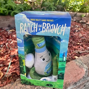 May include: A Hidden Valley Ranch on a Branch plush toy packaged in a rectangular cardboard box. The box features blue sky and green landscape graphics with the text Hidden Valley Ranch Presents Ranch on a Branch. The plush is bottle-shaped.