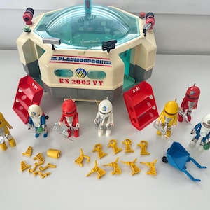 Op de afbeelding: Een Playmobil Space Station set met een beige en teal structuur, rode opslageenheden en verschillende astronautenfiguren in diverse kleuren. De set bevat kleine gele en blauwe accessoires, en de tekst "PLAYMOSPACE" en "RS 2005 VY" is zichtbaar.