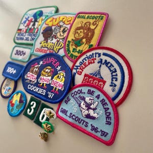 May include: A collection of colorful embroidered patches featuring various themes, including Girl Scouts, cookies, and super heroes. The patches are arranged on a white surface. The text on the patches includes "Girl Scouts", "Cookies '97", "Super", "Team Up", "Marriott's Great America", and "Be Cool, Be a Reader".