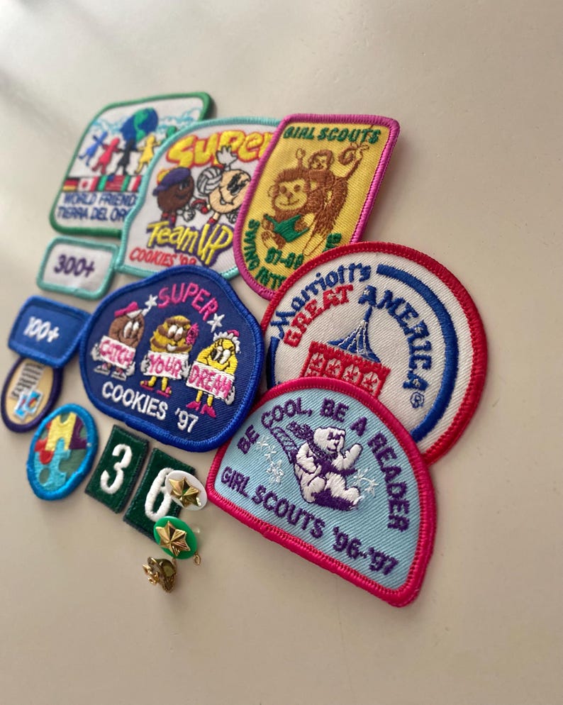 May include: A collection of colorful embroidered patches featuring various themes, including Girl Scouts, cookies, and super heroes. The patches are arranged on a white surface. The text on the patches includes "Girl Scouts", "Cookies '97", "Super", "Team Up", "Marriott's Great America", and "Be Cool, Be a Reader".