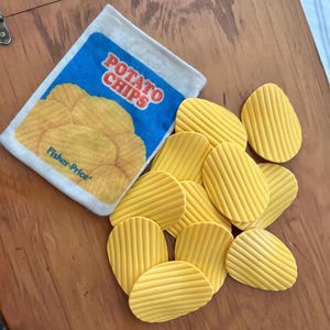 Puede incluir: Un juego de juguetes vintage de Fisher-Price con una bolsa de patatas fritas y varias patatas fritas amarillas y onduladas. La bolsa es azul con las palabras "POTATO CHIPS" en rojo y blanco. El juguete está sobre una superficie de madera.