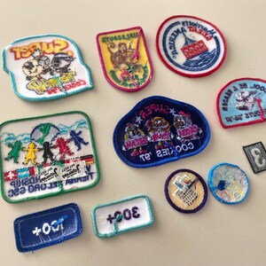 May include: A collection of 11 colorful embroidered patches featuring various designs, including a boat, a group of people, and the words "Cookies", "Sweet", and "Friendship".