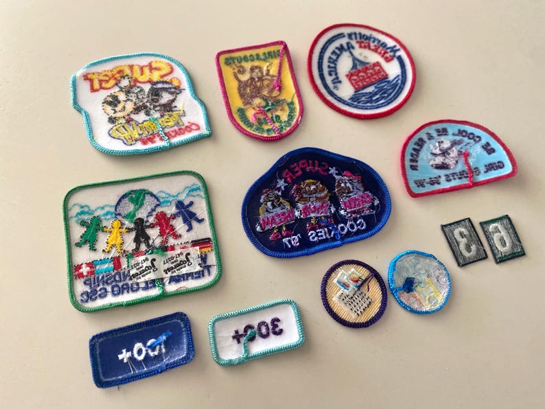 May include: A collection of 11 colorful embroidered patches featuring various designs, including a boat, a group of people, and the words "Cookies", "Sweet", and "Friendship".