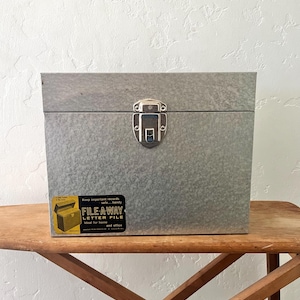 Puede incluir: Una caja de archivos rectangular de acero galvanizado con acabado metálico plateado y un cierre central de metal. Una etiqueta rectangular negra en la parte frontal presenta el texto FILE-A-WAY LETTER FILE, Keep important records safe...handy e Ideal for home and office.