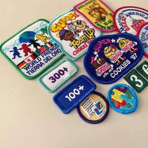 May include: A collection of Girl Scout cookie badges featuring various designs and colors. The badges include "World Friends", "Super Team Up", "Catch Your Dream", "Be a Reader", and "300+".