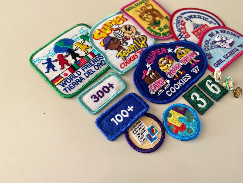 May include: A collection of Girl Scout cookie badges featuring various designs and colors. The badges include "World Friends", "Super Team Up", "Catch Your Dream", "Be a Reader", and "300+".