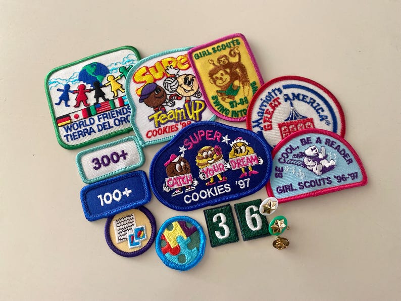 May include: A collection of colorful embroidered patches featuring various Girl Scout themes, including "World Friends", "Super Cookies", "Be Cool, Be a Reader", and "Marriott's Great America".