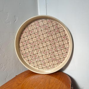 Puede incluir: Bandeja redonda para servir con borde color crema e interior estampado. El interior presenta un diseño repetitivo de pequeñas flores rojas y formas geométricas marrones. La bandeja está hecha de un material claro y se apoya sobre una superficie de madera.