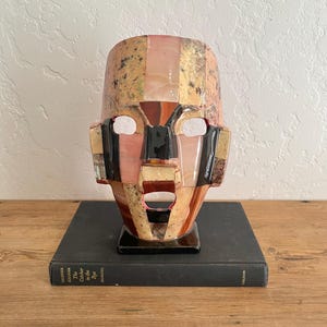 May include: A mosaic mask sculpture with a human face design. The mask features a combination of pink, black, beige, and brown stone pieces. The sculpture is placed on a black book.