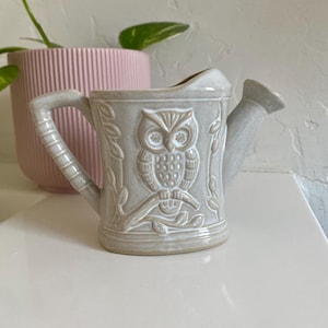 May include: A small, white ceramic watering can with an owl design. The owl is in a beige color and is surrounded by leaves. The watering can has a spout and a handle.