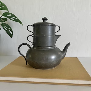 May include: A vintage pewter tea set, featuring a teapot with a curved handle and spout, a sugar bowl, and a creamer. The set is displayed on a tan book, with a green plant in the background. The tea set has a dark, aged appearance.