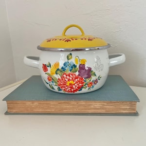 May include: White enamel pot with a yellow lid and floral design. The pot features a red, orange, blue, and purple flower pattern with a silver rim and handles. A butterfly is also depicted on the side.