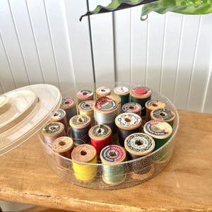 May include: A clear plastic round container filled with spools of colorful thread. The spools are wooden and have various labels with text such as "COATS" and "MERCERIZED SEWING".