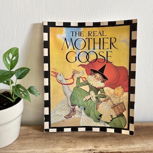 Puede incluir: Un libro infantil vintage titulado "The Real Mother Goose" con una ilustración colorida de una mujer con un vestido verde y una capa roja montando un ganso blanco. Un bebé está sentado en una cesta en la espalda del ganso.