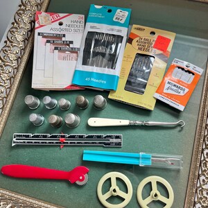 Puede incluir: Una colección de herramientas y suministros de costura vintage, que incluye agujas surtidas, dedales, un abridor de costuras y un calibrador de medición. Los artículos están dispuestos sobre una superficie verde dentro de una bandeja decorativa con marco dorado. Se ven varios paquetes de agujas.