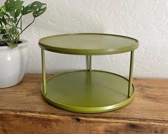 Puede incluir: Un organizador de almacenamiento circular de dos niveles en color verde oliva, con una base plana y un estante superior a juego sostenido por tres pilares verticales delgados. La estructura está hecha de un material sólido y liso.
