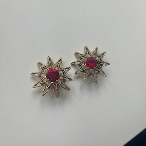 May include: A pair of gold-toned starburst earrings, each featuring a vibrant red gemstone center. The earrings have a delicate, openwork design, creating a vintage aesthetic. The earrings are displayed on a white surface.