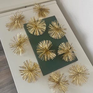 Pode incluir: Uma coleção de ornamentos decorativos dourados em forma de estrela, dispostos sobre uma superfície branca, alguns colocados ao redor de um livro verde escuro fechado. Os ornamentos têm um design radial e pontiagudo, criando uma exibição festiva.