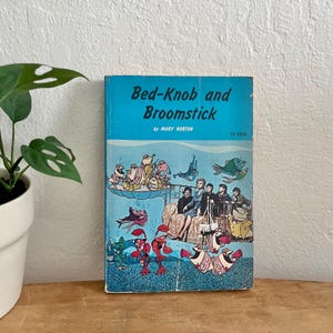 Könnte beinhalten: Ein Vintage-Exemplar von "Bed-Knob and Broomstick" von Mary Norton. Das Buchcover zeigt eine lebendige Illustration mit blauem Hintergrund und skurrilen Figuren. Der Titel ist weiß, darunter der Name des Autors.
