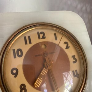 May include: A vintage General Electric alarm clock with a white face and gold-colored hands. The clock has a brown center with a gold-colored design. The clock is set to 12:00.
