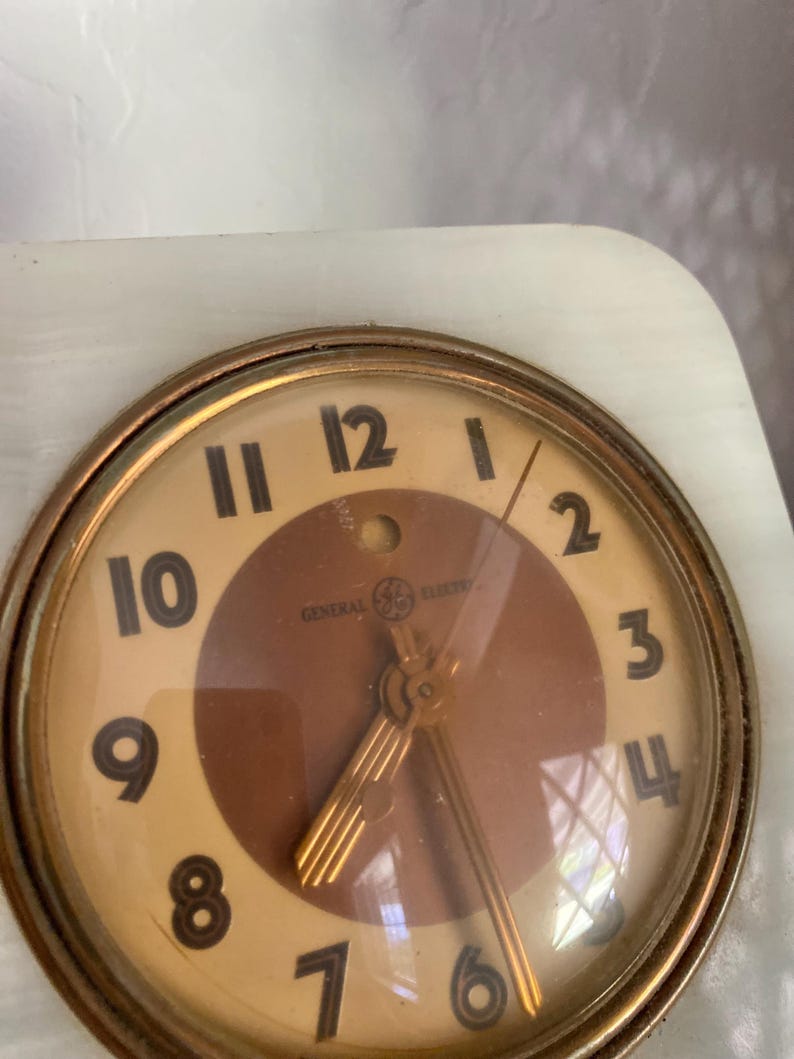 May include: A vintage General Electric alarm clock with a white face and gold-colored hands. The clock has a brown center with a gold-colored design. The clock is set to 12:00.