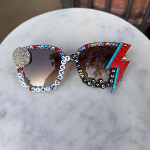 Può includere: Occhiali da sole con lenti sfumate marroni e montatura decorata con strass colorati. La lente sinistra presenta un accento glitter circolare, mentre la destra un fulmine rosso. Perline con lettere compongono la scritta THIS AINT NO PARTY THIS AINT NO DISCO.