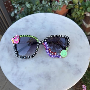 Può includere: Occhiali da sole neri con accenti decorativi di perline. Il lato sinistro presenta un cuore rosa e le parole "AFTER ALL I BE". Il lato destro ha le parole "ONE THAT MATTERS" e una faccina verde sorridente. Le montature sono impreziosite da perline verdi e viola.