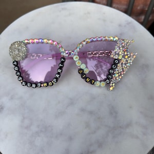 Può includere: Occhiali da sole viola chiaro con montatura decorata da strass iridescenti, un grande accento circolare in cristallo e una forma a fulmine. Perline con lettere nere e pastello compongono la frase THIS AINT NO PARTY THIS AINT NO DISCO.