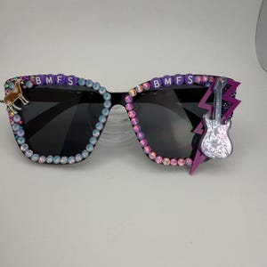 Puede incluir: Gafas de sol negras con adornos decorativos. Las monturas están adornadas con pedrería iridiscente y las letras "BMFS" a cada lado. Una pequeña guitarra y un rayo acentúan el lado derecho, y un pequeño dije de perro está a la izquierda.