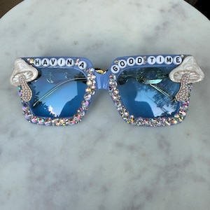 Può includere: Occhiali da sole con montatura quadrata azzurra e lenti blu, decorati con strass iridescenti lungo i bordi inferiori. Le montature sono arricchite da ciondoli a forma di fungo bianco sugli angoli esterni e perline con lettere che compongono HAVIN A GOOD TIME.