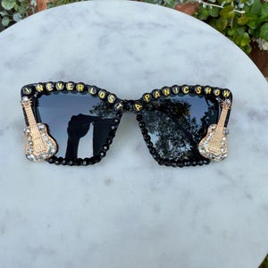 May include: Black sunglasses with the text "I NEVER LOST A PANIC SHOW" in gold and black beads. The sunglasses have a black frame with rhinestones and gold guitar accents on each side.