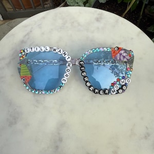 May include: Clear-framed sunglasses with blue lenses. The frames are embellished with colorful rhinestones and beads spelling out "STILL WANT MORE" and "FISHWATER MOMO". Decorative patches and charms adorn the corners, adding a unique, artistic flair.