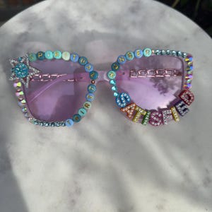 May include: Clear-framed sunglasses with purple lenses, embellished with colorful beads spelling "SPIRITUAL CHANGE" and "SATISFIED". The frames are also adorned with rhinestones and a star-shaped charm.