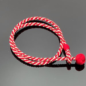 May include: A red and white braided cord with a decorative red ball at the end. The cord is looped in a circular shape. The cord has a twisted pattern with red, white, and tan threads.