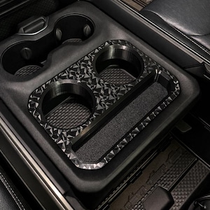 May include: A black automotive center console cup holder insert featuring a geometric, faceted pattern on the top surface. The insert includes two circular cup holders and a rectangular storage tray, designed to fit into the existing vehicle console.