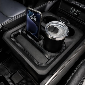 May include: A black carbon fiber patterned center console organizer tray for a vehicle, featuring a dedicated upright slot for a smartphone and a circular cutout for a beverage tumbler, designed to fit securely within the existing cup holder area.