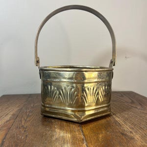 May include: A vintage brass bucket with a curved handle. The bucket has a square base and is decorated with a repeating fan-like pattern. The brass has a warm, aged patina, and the bucket sits on a wooden surface.