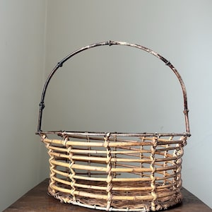 May include: A woven wicker basket with a brown metal handle. The basket is oval shaped and has a natural, light brown color.
