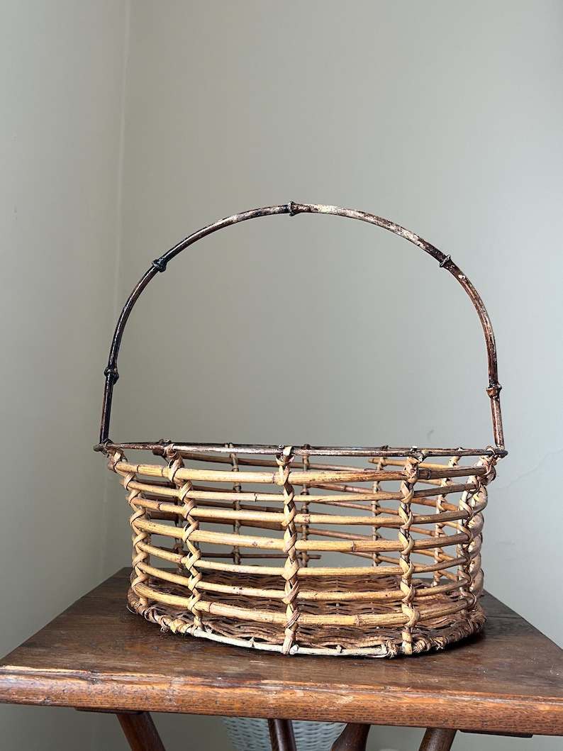 May include: A woven wicker basket with a brown metal handle. The basket is oval shaped and has a natural, light brown color.