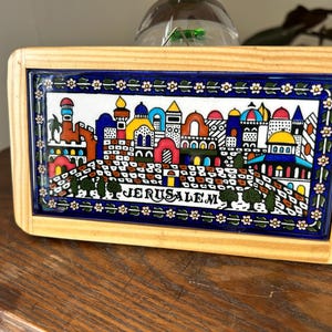 May include: A wooden framed tile with a colorful illustration of a cityscape with the word "JERUSALEM" in the center. The tile is decorated with a blue and white border with white flowers.