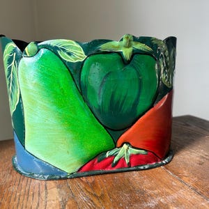 May include: A hand-painted, cylindrical container featuring a vibrant vegetable design. The artwork includes a green bell pepper, a tomato, and a pear, with green and red hues. The container sits on a wooden surface.