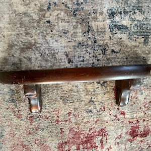 May include: A dark brown wooden shelf with curved edges and decorative scrollwork on the ends. The shelf is mounted on a wall.