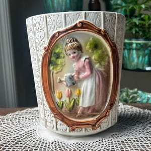 May include: Cream ceramic planter with a raised relief design. An oval frame shows a woman watering flowers. The planter has a textured surface and a brown border around the oval. The theme is vintage decor.
