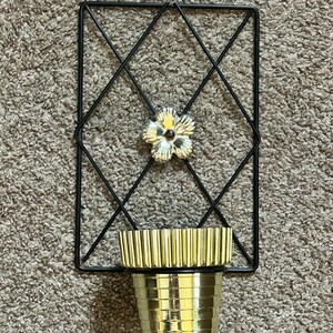 May include: A black metal wall sconce with a diamond pattern and a gold-colored flower accent. The sconce features a gold-colored cup-shaped holder. The sconce is designed to hold a small plant or candle.