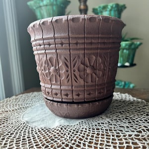 May include: A brown, carved ceramic flower pot with a matching saucer. The pot features a detailed floral and geometric design. The pot is sitting on a white doily on a wooden table. Other green pots are in the background.