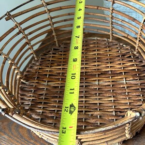 May include: A woven wicker basket with a metal frame. The basket is brown and has a circular shape. A measuring tape is placed next to the basket, showing a measurement of 13 inches.